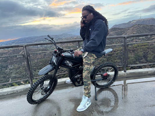 Man in camo pants and sneakers sitting on a black electric bike outdoors with scenic mountain background at sunset.