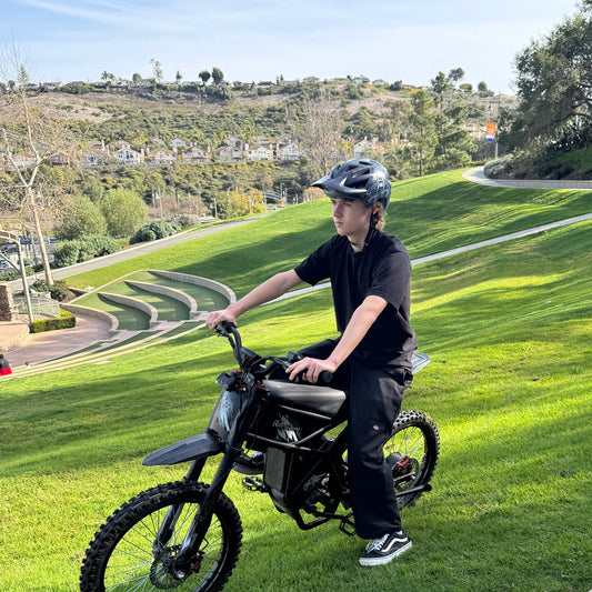 A rider in a black t-shirt and helmet sitting on a black Riding'times GT73 E-Motorbike on a lush green grass hill overlooking a valley with houses