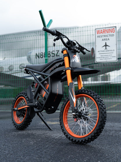 GT54 electric mini motorbike with orange accents and off-road tires parked near airfield fence