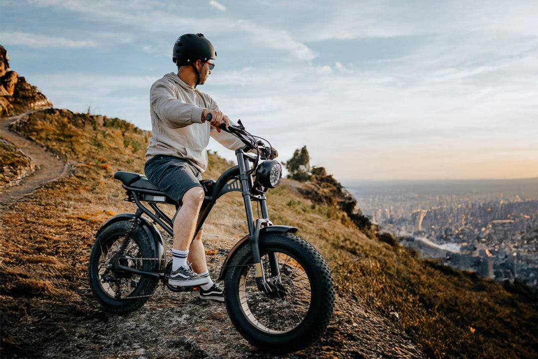 Black rugged E-Bike with wide all-terrain tires, suspension fork, and round headlight on a dirt trail with a rider wearing helmet