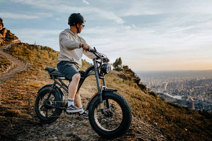 Black rugged E-Bike with wide all-terrain tires, suspension fork, and round headlight on a dirt trail with a rider wearing helmet
