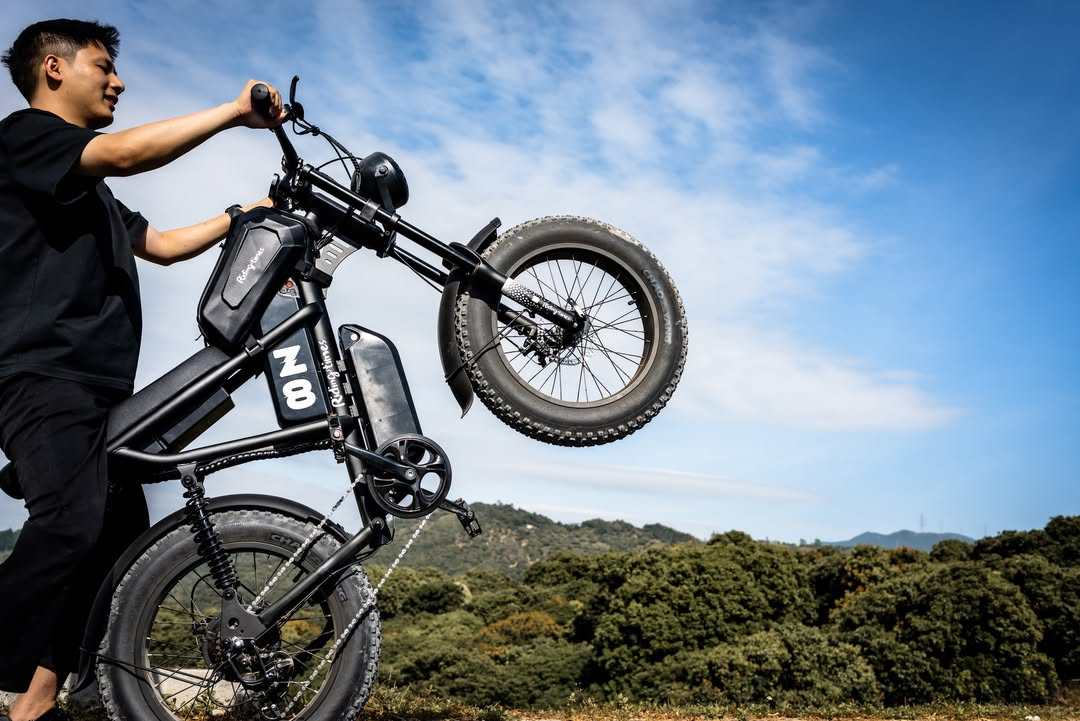 Black matte E-Bike with thick 20-inch all-terrain tires, sturdy frame, and front suspension being lifted by a person